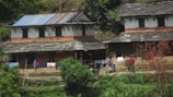 A rustic rural setting with two traditional houses made of stone and clay, featuring slate roofs. Laundry hangs drying on a line in front of the houses. Two people are visible, with one carrying a bundle on their back. The surroundings are green with vegetation and a few trees.