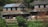 A rustic rural setting with two traditional houses made of stone and clay, featuring slate roofs. Laundry hangs drying on a line in front of the houses. Two people are visible, with one carrying a bundle on their back. The surroundings are green with vegetation and a few trees.