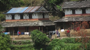 A rustic rural setting with two traditional houses made of stone and clay, featuring slate roofs. Laundry hangs drying on a line in front of the houses. Two people are visible, with one carrying a bundle on their back. The surroundings are green with vegetation and a few trees.
