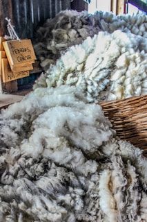 A close-up view of piles of raw wool in neutral shades, arranged in wooden or wicker containers. Signage with handwritten labels indicating different categories of wool is visible.