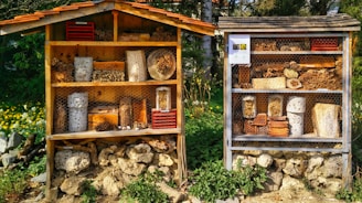 Two wooden structures resembling insect hotels are placed side by side outdoors. Each structure contains various natural materials like logs, sticks, and bricks with holes to provide shelter for insects. The structures are surrounded by lush greenery and flowers, creating a harmonious blend with the natural environment.