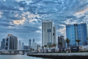 A cityscape featuring modern high-rise buildings beneath a dramatic, cloudy sky. The skyline includes a prominent skyscraper with the label 'DAMAC' alongside other tall structures. Palm trees line the waterfront, and a bridge crosses over a body of water in the foreground.