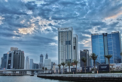 A cityscape featuring modern high-rise buildings beneath a dramatic, cloudy sky. The skyline includes a prominent skyscraper with the label 'DAMAC' alongside other tall structures. Palm trees line the waterfront, and a bridge crosses over a body of water in the foreground.