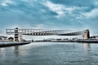 A modern pedestrian bridge spans across the water, featuring a sleek, architectural design with angular, metallic structures and glass facades. The bridge is elevated, supported by vertical pillars, and stretches across the river, connecting two urban areas. The sky is overcast, with a mix of dark and light clouds, creating a dramatic atmosphere.