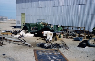 A classic green car is parked outdoors near a corrugated metal building. Two people appear to be working underneath the vehicle, which is propped up. Various tools and equipment, including pipes and metal frames, are scattered around. The surrounding environment seems industrial with a sandy ground and a distant view of a fenced structure.