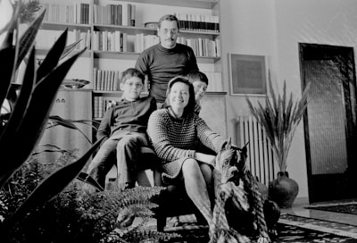 A family of four poses in a living room, with a dog seated in the foreground. The room is decorated with potted plants, bookshelves filled with books, and a framed picture on the wall. The family members are dressed in casual clothing and are seated around an armchair.