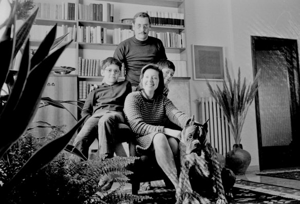 A family of four poses in a living room, with a dog seated in the foreground. The room is decorated with potted plants, bookshelves filled with books, and a framed picture on the wall. The family members are dressed in casual clothing and are seated around an armchair.