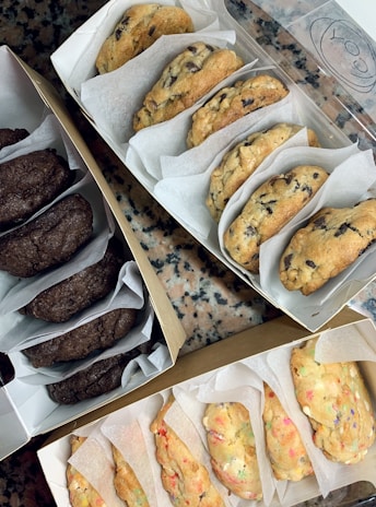 Three open boxes containing different varieties of cookies, such as chocolate chip, double chocolate, and funfetti, placed on a speckled countertop.