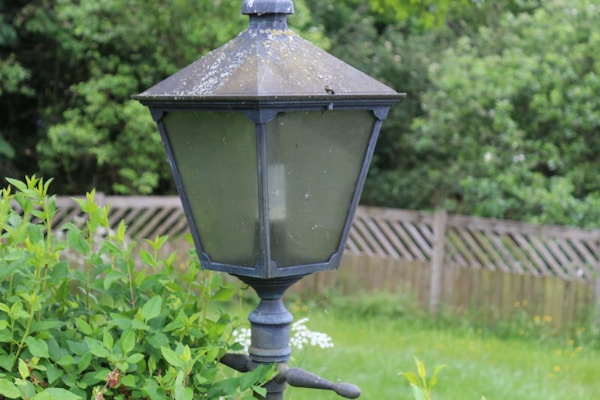 An old, weathered metal lantern post stands surrounded by lush green foliage. The lantern features a dusty, inclined top with glass panels, showing signs of age and exposure to the elements. In the background, a wooden fence and dense green trees create a serene garden atmosphere.