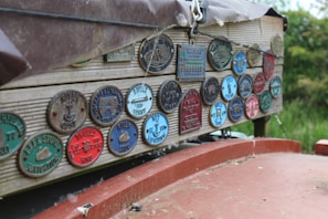 A collection of colorful, vintage-looking plaques are displayed on a wooden surface. Each plaque is circular or rectangular, featuring embossed lettering and images specific to different associations or locations, likely related to navigation or waterways. The plaques have a weathered and rustic charm, and their varied colors stand out against the wood. The background shows some greenery, indicating an outdoor setting.