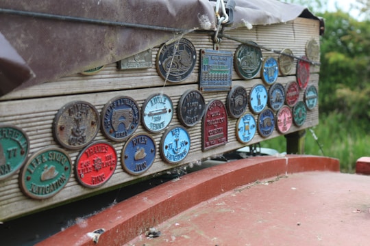 A collection of colorful, vintage-looking plaques are displayed on a wooden surface. Each plaque is circular or rectangular, featuring embossed lettering and images specific to different associations or locations, likely related to navigation or waterways. The plaques have a weathered and rustic charm, and their varied colors stand out against the wood. The background shows some greenery, indicating an outdoor setting.