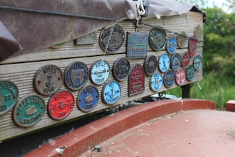 A collection of colorful, vintage-looking plaques are displayed on a wooden surface. Each plaque is circular or rectangular, featuring embossed lettering and images specific to different associations or locations, likely related to navigation or waterways. The plaques have a weathered and rustic charm, and their varied colors stand out against the wood. The background shows some greenery, indicating an outdoor setting.