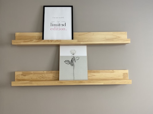 A minimalist wall display features two wooden shelves mounted on a plain gray wall. The top shelf holds a framed print with text saying 'i’m not weird, i’m limited edition.' Below it, a second piece of artwork shows a simple drawing of a rose in black and white.