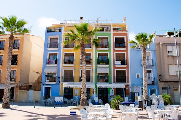A vibrant street scene featuring a series of colorful, multi-story buildings with balconies. These buildings are painted in bright shades of blue, green, and orange. Palm trees line the street, adding a tropical atmosphere. In the foreground, there are empty tables and chairs arranged in an outdoor seating area, likely belonging to a cafe or restaurant.