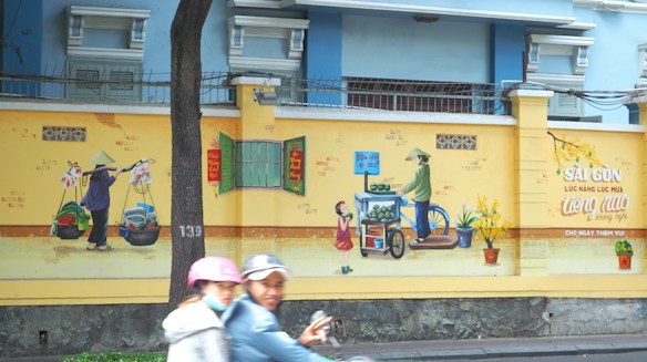 A yellow wall features vibrant street art depicting traditional Vietnamese street vendors. One vendor wears a conical hat and carries produce on a shoulder pole, while another sells watermelons from a cart. There is green foliage in flowerpots placed on the ground. Two people on a motorbike are passing by in the foreground. Above the artwork, a blue and white building is partially visible with barred windows.