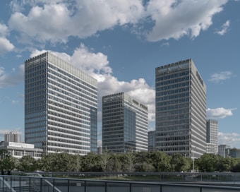 Three modern office buildings with glass exteriors rise against a blue sky scattered with white clouds. Below the buildings, a line of green trees adds a touch of nature to the urban landscape. A walkway with metal railings crosses in the foreground, leading the eye into the city scene.