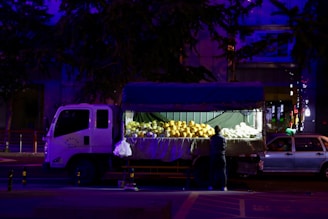 A truck with an open back is parked on the street at night. The back of the truck is filled with large fruits, possibly melons, under bright artificial lighting. A person stands beside the truck, seemingly arranging or selecting the fruits.