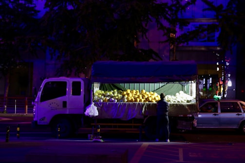 A truck with an open back is parked on the street at night. The back of the truck is filled with large fruits, possibly melons, under bright artificial lighting. A person stands beside the truck, seemingly arranging or selecting the fruits.