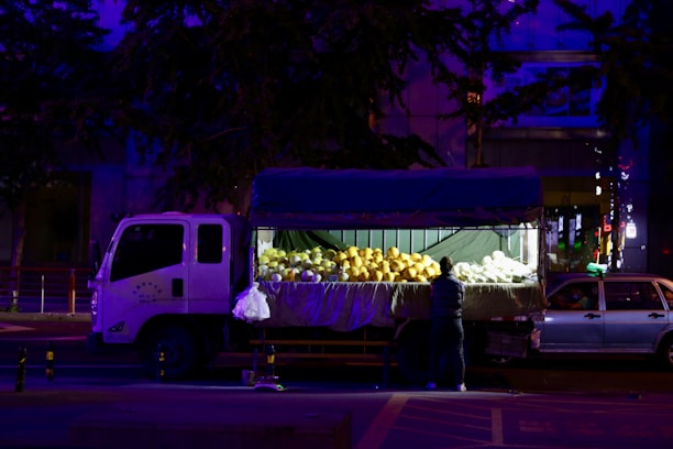 A truck with an open back is parked on the street at night. The back of the truck is filled with large fruits, possibly melons, under bright artificial lighting. A person stands beside the truck, seemingly arranging or selecting the fruits.