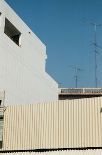 An urban landscape features a minimalist architectural design with white and cream-colored buildings. Multiple television antennas rise against a clear blue sky, adding a sense of quiet simplicity.