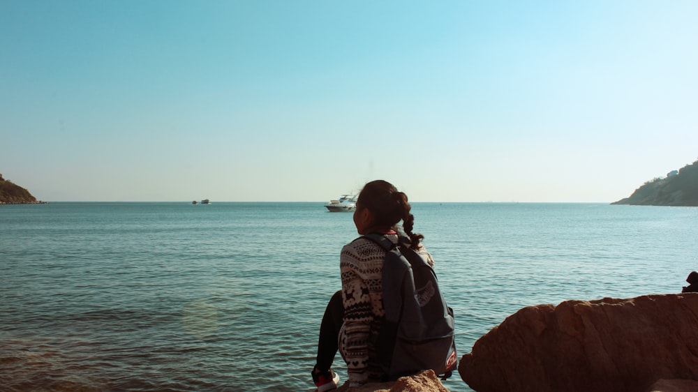 man in black and gray jacket sitting on brown rock formation near body of water during