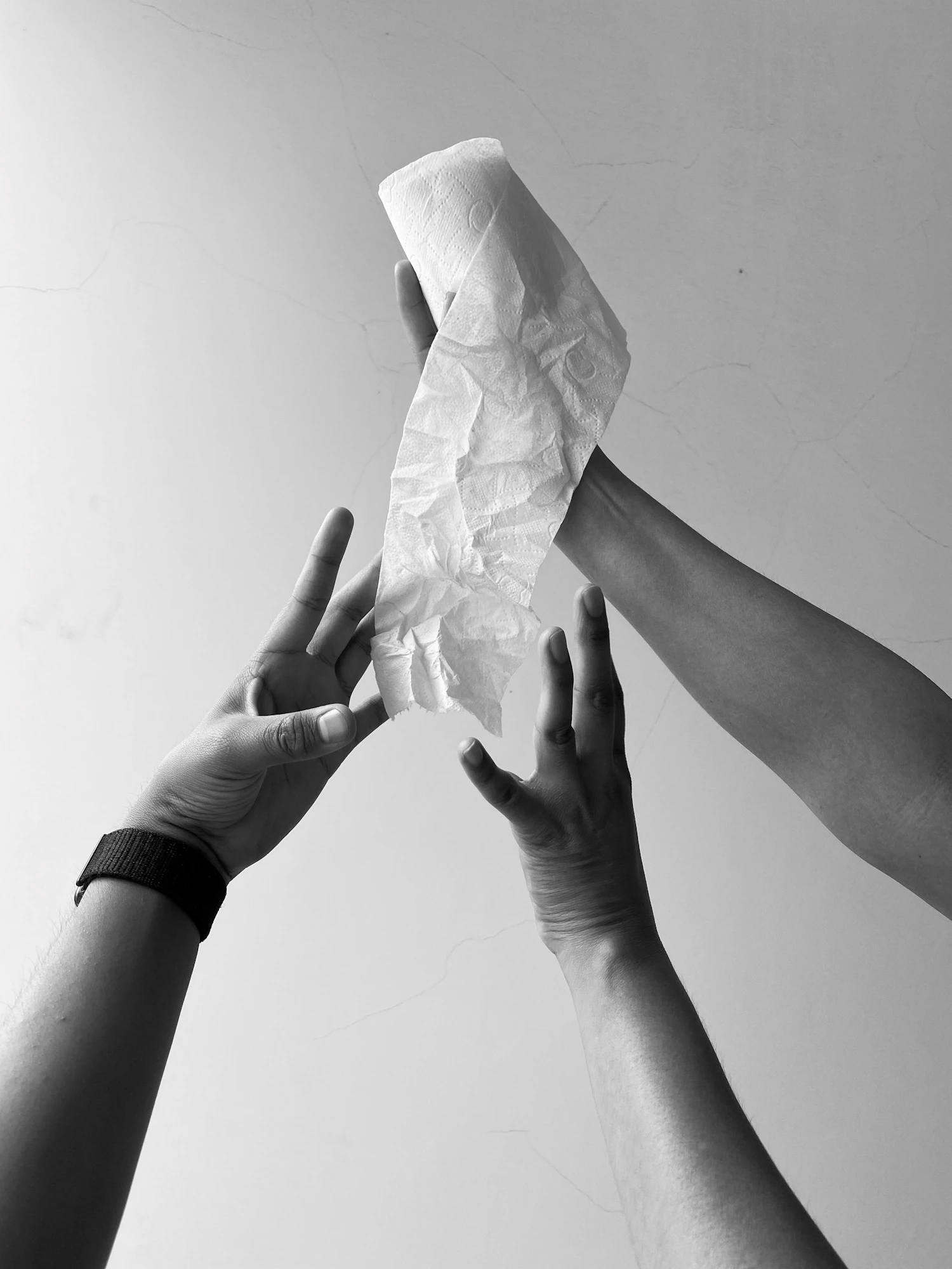 Three hands reaching up to hold a single crumpled sheet of white tissue against a pale wall