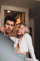 A man and a woman are posing indoors. The man wears a gray sweater, while the woman has on a white cardigan and jeans. Both have relaxed yet confident expressions.