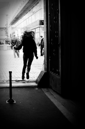 A person is captured mid-jump while exiting a dark tunnel or passageway into a brightly lit street. In the background, several other pedestrians walk along a cobblestone road lined with buildings featuring large windows and an architectural awning.