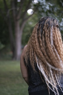A person with long, curly hair standing outdoors, surrounded by greenery with trees in the background. The focus is on the back of the person, emphasizing their hairstyle.
