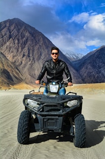A person wearing a black leather jacket and sunglasses is riding an all-terrain vehicle (ATV) on a sandy terrain. The background features rugged mountains under a partially cloudy blue sky.
