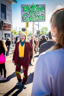 A person dressed in a colorful costume with makeup holds a sign advocating for justice in a crowd of people at a daytime gathering. The scene is lively, with numerous individuals in the background wearing casual clothing, and a bright sun casting shadows on the ground.