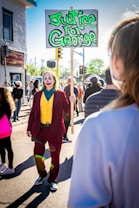 A person dressed in a colorful costume with makeup holds a sign advocating for justice in a crowd of people at a daytime gathering. The scene is lively, with numerous individuals in the background wearing casual clothing, and a bright sun casting shadows on the ground.