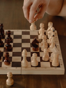 A hand is moving a white chess piece on a wooden chessboard. The board is set up for a game, showing both light and dark wooden pieces in various positions. The focus is on the movement of the chess piece, suggesting an active gameplay.
