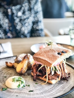 A delicious sandwich filled with shredded meat and fresh vegetables, served on a plate with roasted potato wedges and a dollop of creamy sauce garnished with herbs. The setting appears to be casual dining, with a bokeh effect in the background indicating a vibrant, lively atmosphere.