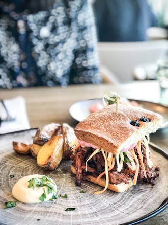A delicious sandwich filled with shredded meat and fresh vegetables, served on a plate with roasted potato wedges and a dollop of creamy sauce garnished with herbs. The setting appears to be casual dining, with a bokeh effect in the background indicating a vibrant, lively atmosphere.
