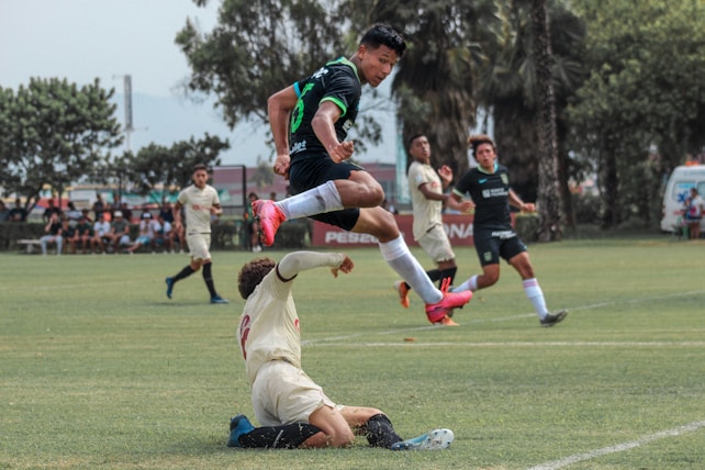 A dynamic scene of a soccer match with a player in a green jersey leaping over another player in a white jersey. The green jerseyed player is airborne, while the white jerseyed player is on the ground attempting to tackle. Other players and spectators are visible in the background.