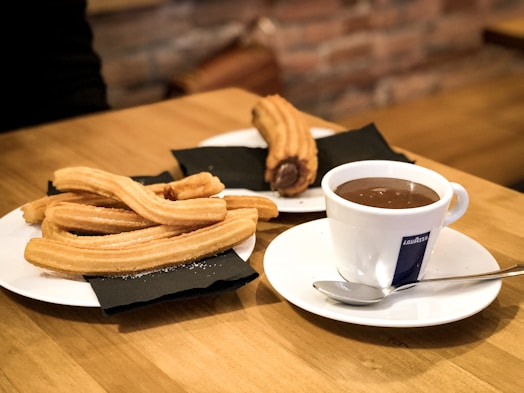 A wooden table setting includes a plate of churros and a cup of chocolate. The churros are golden and sugar-coated, lying on a white plate with a black napkin beneath them. Nearby, a coffee cup filled with hot chocolate sits on a saucer, accompanied by a spoon.