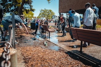 A group of people are actively participating in cleaning an outdoor area. Some are using brooms and shovels to clear debris and water from a pathway, while others work together to manage the surroundings. The scene is set against a backdrop of trees and a building, with bright sunlight highlighting the activity.
