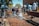 A group of people are actively participating in cleaning an outdoor area. Some are using brooms and shovels to clear debris and water from a pathway, while others work together to manage the surroundings. The scene is set against a backdrop of trees and a building, with bright sunlight highlighting the activity.