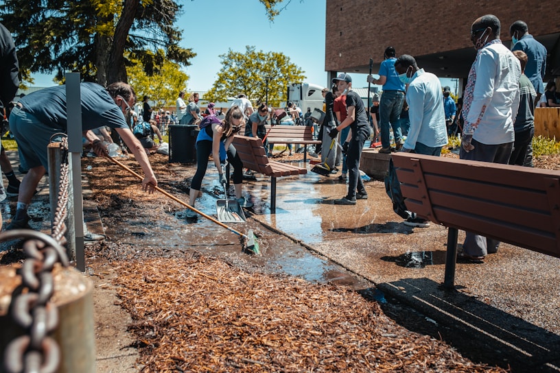 A group of people are actively participating in cleaning an outdoor area. Some are using brooms and shovels to clear debris and water from a pathway, while others work together to manage the surroundings. The scene is set against a backdrop of trees and a building, with bright sunlight highlighting the activity.
