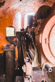 A detailed view of what appears to be the interior of an old, rusted industrial machine or boiler. The image showcases intricate mechanical components including large gears and pipes. The surfaces are covered with rust and patches of orange paint, giving the machinery a rugged and weathered appearance.