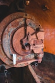 A close-up of an old, rusted industrial machine part with visible corrosion. The metal surface displays various shades of rust and signs of aging. There is a white cylindrical object attached to the machinery, contrasting with the rusty texture. The background features an orange-brown hue, adding a warm tone to the scene.