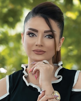A woman gazes confidently at the camera, her hand resting thoughtfully under her chin. Her hair is elegantly styled, and she wears makeup that accentuates her features. She is dressed in a black top with white trim and adorned with jewelry, including a striking ring and a watch. The background is a soft focus of greenery, suggesting an outdoor setting.