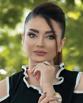 A woman gazes confidently at the camera, her hand resting thoughtfully under her chin. Her hair is elegantly styled, and she wears makeup that accentuates her features. She is dressed in a black top with white trim and adorned with jewelry, including a striking ring and a watch. The background is a soft focus of greenery, suggesting an outdoor setting.