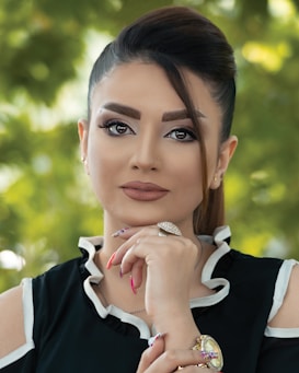 A woman gazes confidently at the camera, her hand resting thoughtfully under her chin. Her hair is elegantly styled, and she wears makeup that accentuates her features. She is dressed in a black top with white trim and adorned with jewelry, including a striking ring and a watch. The background is a soft focus of greenery, suggesting an outdoor setting.