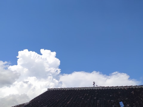 Blue sky with large, fluffy white clouds and the edge of a dark shingled roof at the bottom of the image.