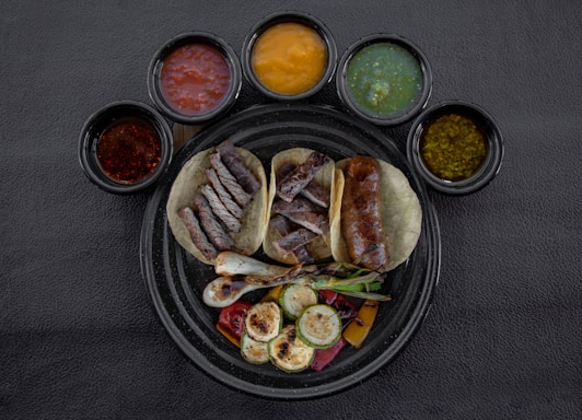 A round black plate contains three tacos filled with grilled meats and vegetables, surrounded by six small bowls of various colored sauces. The tacos are accompanied by grilled onions and slices of zucchini, and the sauces include red, yellow, green, and brown varieties.