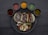 A round black plate contains three tacos filled with grilled meats and vegetables, surrounded by six small bowls of various colored sauces. The tacos are accompanied by grilled onions and slices of zucchini, and the sauces include red, yellow, green, and brown varieties.
