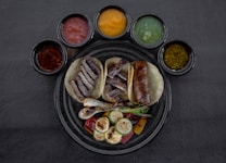 A round black plate contains three tacos filled with grilled meats and vegetables, surrounded by six small bowls of various colored sauces. The tacos are accompanied by grilled onions and slices of zucchini, and the sauces include red, yellow, green, and brown varieties.