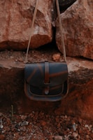 A leather bag with a flap closure is hanging against a backdrop of large, rugged rocks. The bag has a simple design and a natural brown tone, blending with the earthy surroundings of rocky, uneven ground.
